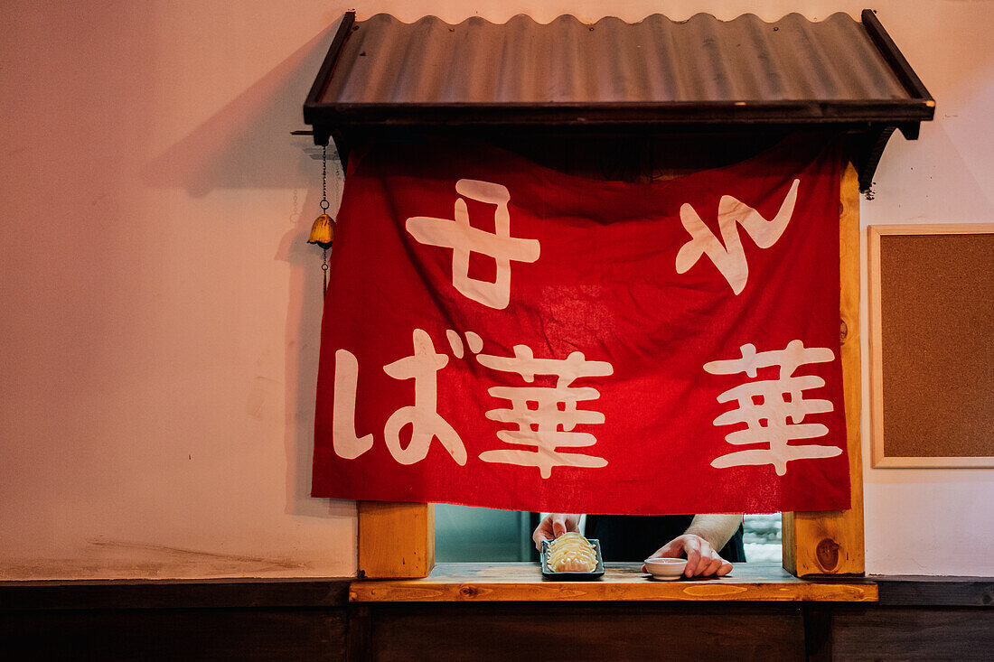 Crop person putting Asian dish with sauce on wooden board with red cloth on window in cafe