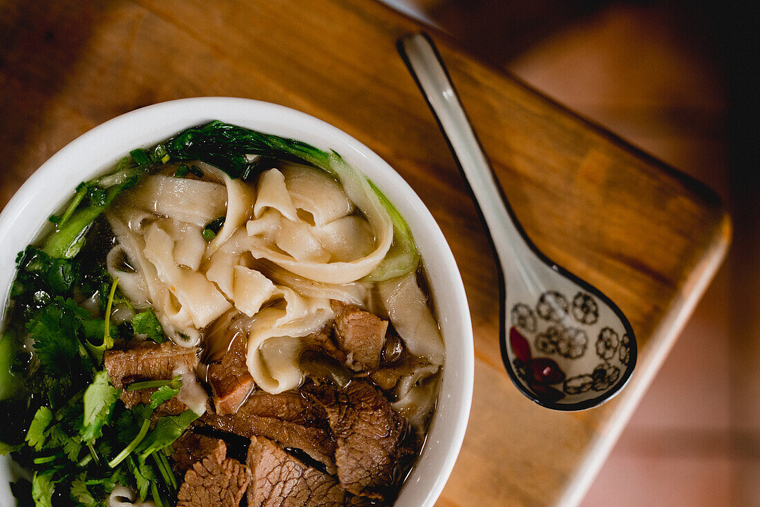 Von oben große weiße Keramikschüssel mit Rindfleisch-Ramen-Suppe und buntem Löffel auf Holztisch platziert