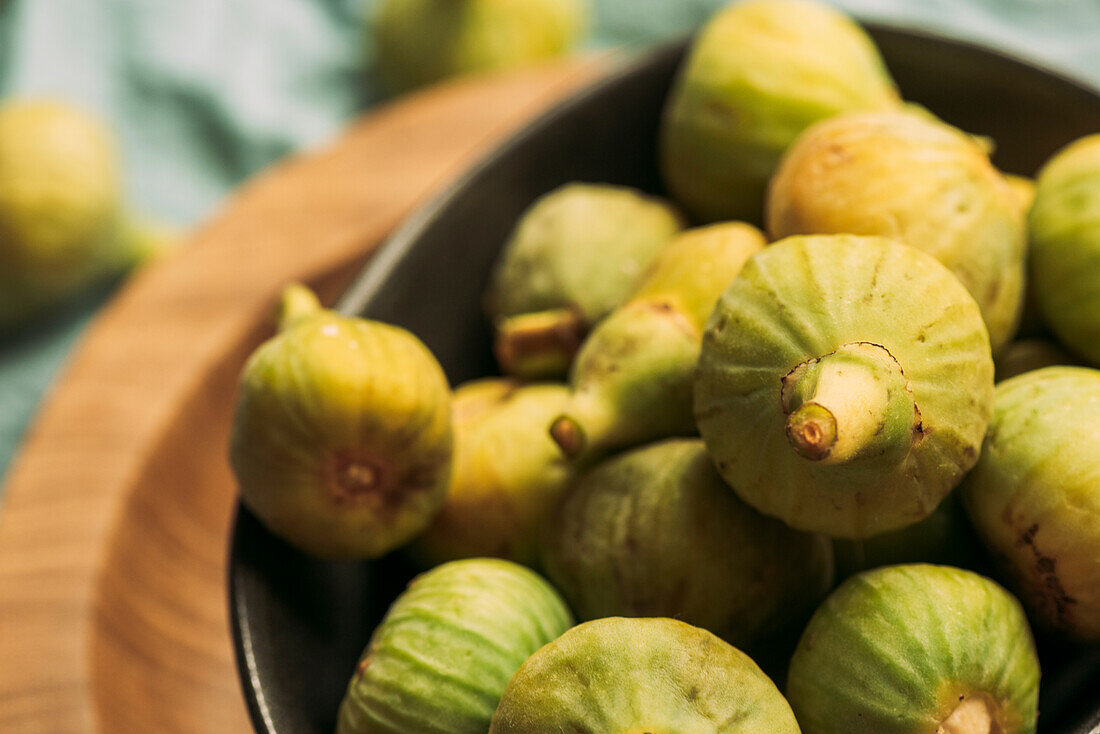 Frische und reife süße grüne Feigen, in einer schwarzen Schale auf dem Holzteller serviert auf dem Tisch mit blauer Tischdecke, Bio-Obst der Saison. Auch bekannt als reife weiße Feigen