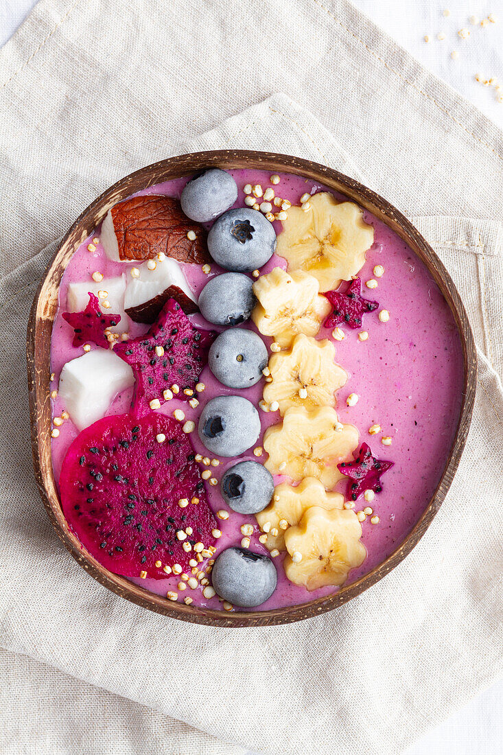 Blick von oben auf eine Schüssel mit einem leckeren Smoothie neben reifen Bananen- und Drachenfruchtscheiben mit Heidelbeeren zum Frühstück