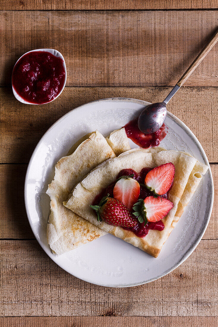 Draufsicht auf leckere Crêpes mit süßer Erdbeermarmelade auf einem Teller neben einem Löffel auf einem Holztisch