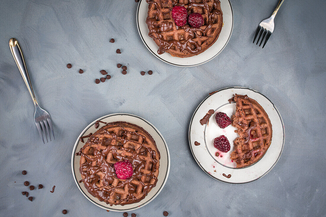 Von oben: leckere süße Waffeln mit Schokoladensauce und Himbeeren, serviert auf einem Tisch mit Schokoladendrops in einer hellen Küche