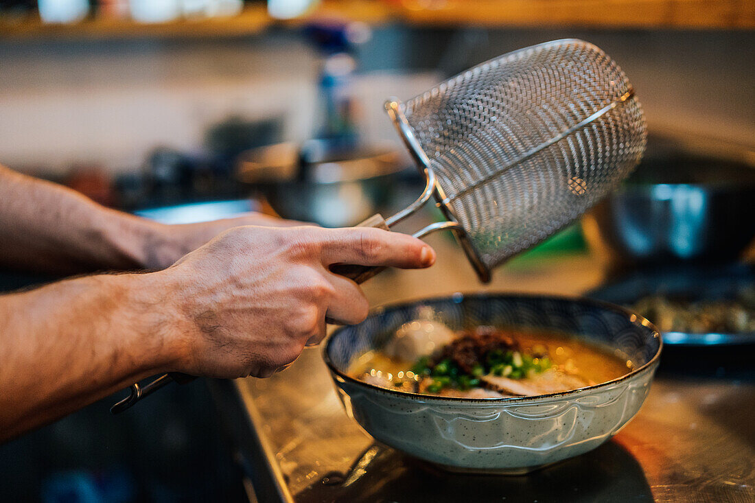 Blick von oben auf einen gesichtslosen Koch, der in einem Café traditionelle Ramen mit Kräutern und Gemüse zubereitet