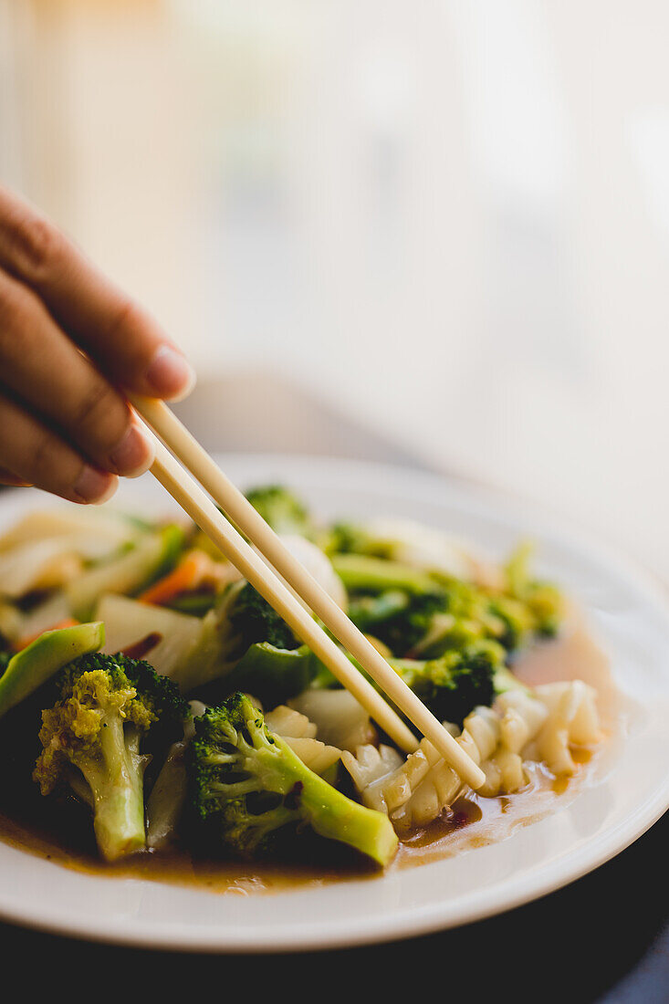 Hand with chopsticks lifting piece from white ceramic plate with broccoli and squid meal