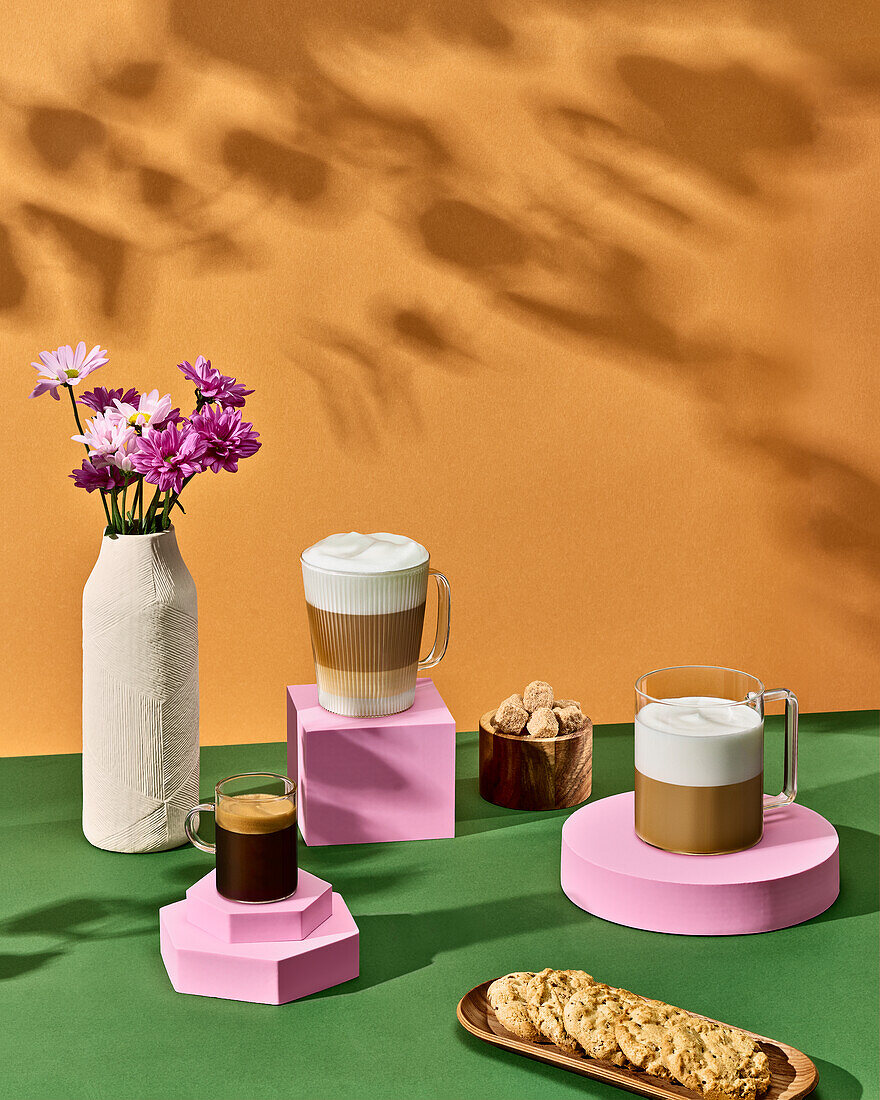 Composition of glass cups with aromatic coffee placed against raisin cookies and vase with flowers on green table