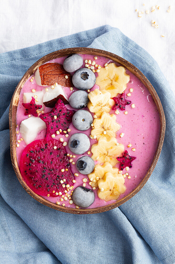 Blick von oben auf eine Schüssel mit einem leckeren Smoothie neben reifen Bananen- und Drachenfruchtscheiben mit Heidelbeeren zum Frühstück