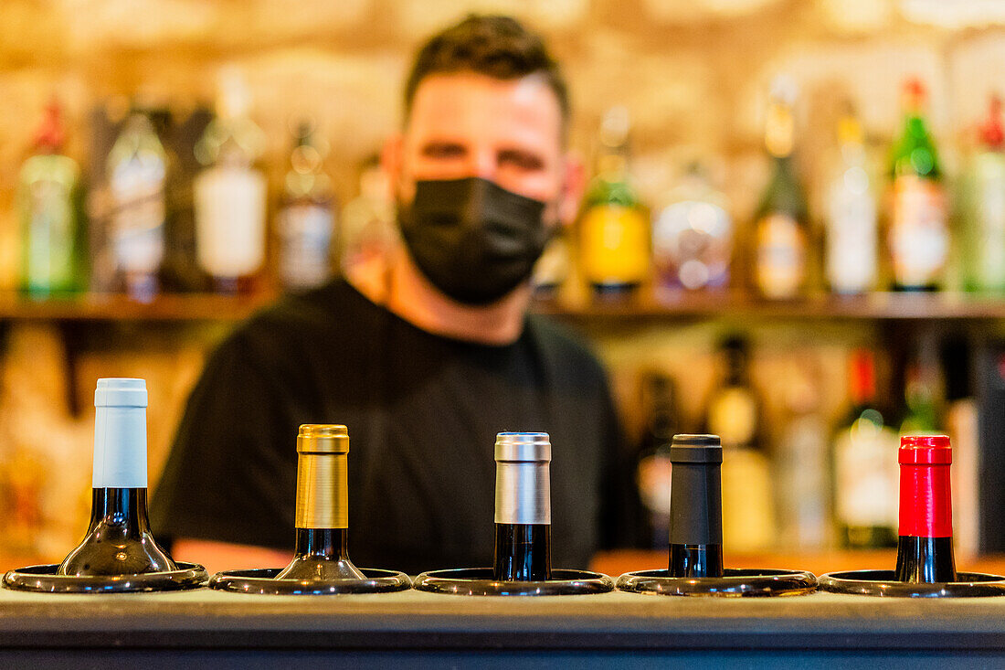 Unscharfer Barmann mit steriler Maske steht am Bartresen, während er in einem Restaurant während des Coronavirus arbeitet