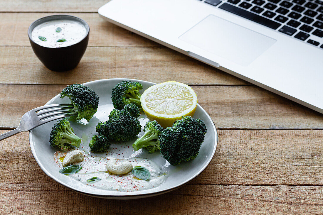 Yummy cooked broccoli with lemon slice and cashew nuts near bowl with white sauce and netbook on wooden table