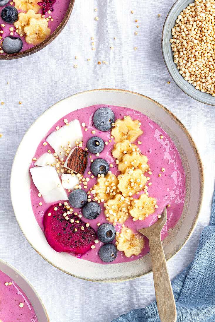 Blick von oben auf eine Schüssel mit einem leckeren Smoothie neben reifen Bananen- und Drachenfruchtscheiben mit Heidelbeeren zum Frühstück