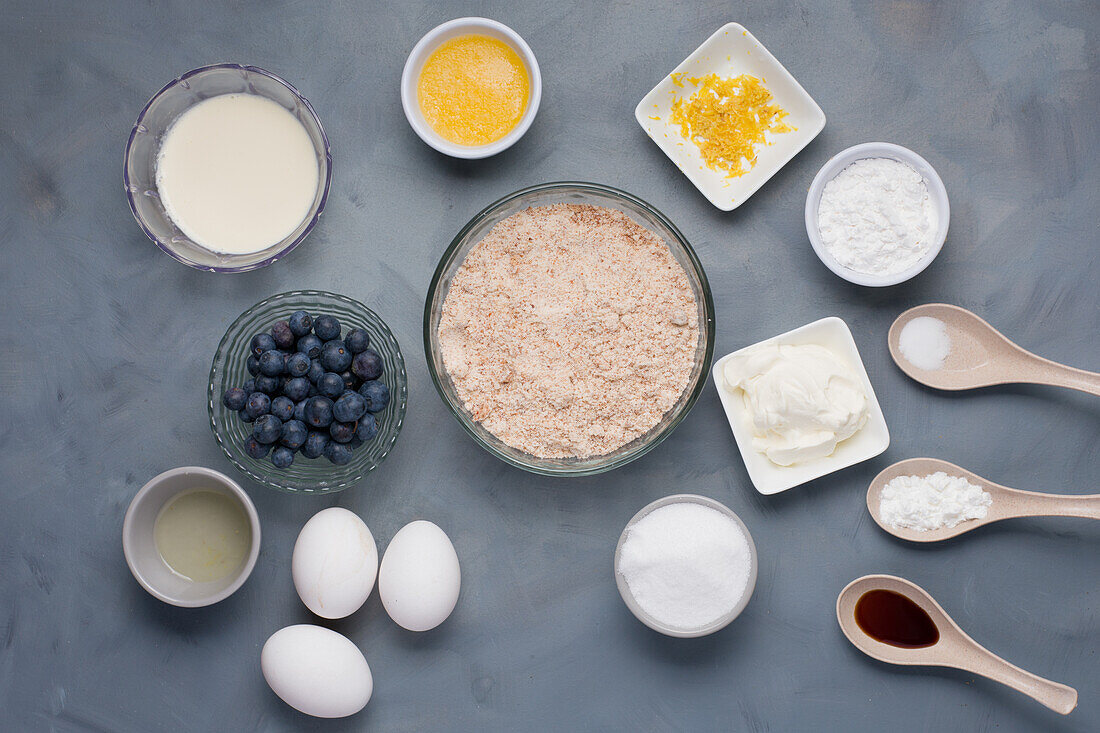 Draufsicht auf verschiedene Zutaten, darunter Milch, geschmolzene Butter, neben Schüsseln mit Zitronenschale, Mehl, Zucker, Salz, neben saurer Sahne, Soda, Öl und Blaubeeren, Eiern und Aromastoffen für ein Gebäckrezept