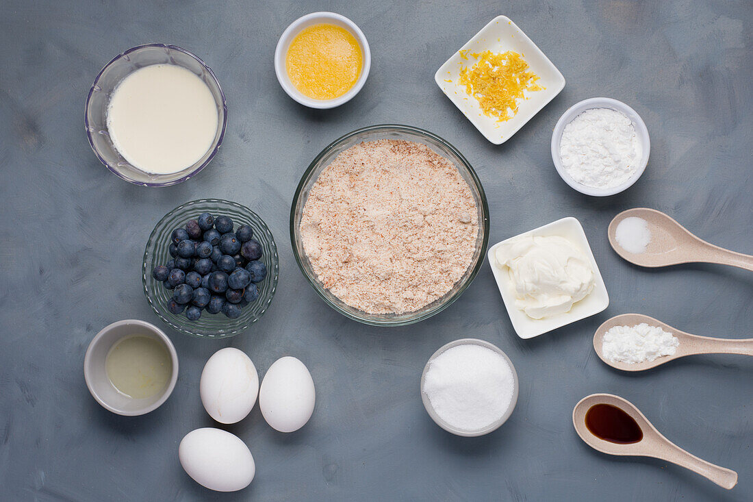 Top view of different ingredients including milk melted butter near bowls of lemon zest flour sugar salt near sour cream soda oil and blueberries eggs with flavoring for pastry recipe