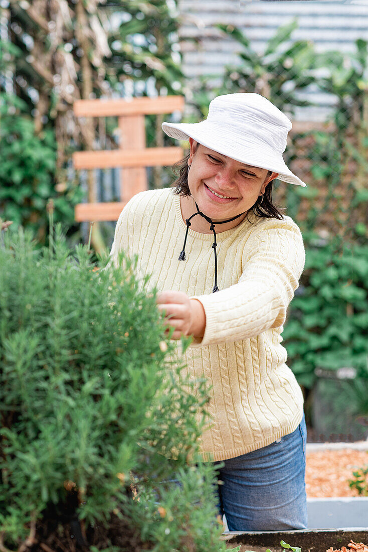 Positive Gärtnerin mit Hut, Pullover und Jeans, die grüne Zweige einer Pflanze im Gartenbeet auf einem Bauernhof berührt