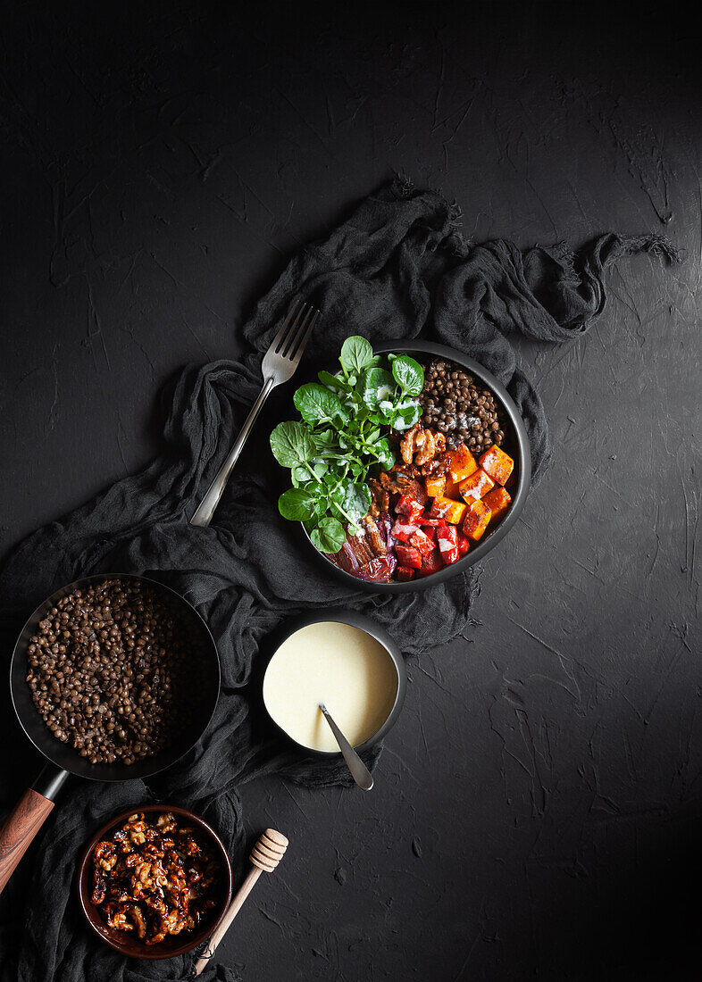 Draufsicht auf Eintopf mit Linsen neben Salat mit Kürbis und Paprika, dekoriert mit grünen Basilikumblättern auf schwarzem Hintergrund neben Salatdressing