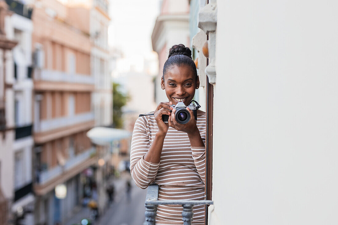 Zufriedene ethnische Frau in gestreifter Kleidung mit professionellem Fotogerät schaut tagsüber auf dem Balkon in die Kamera