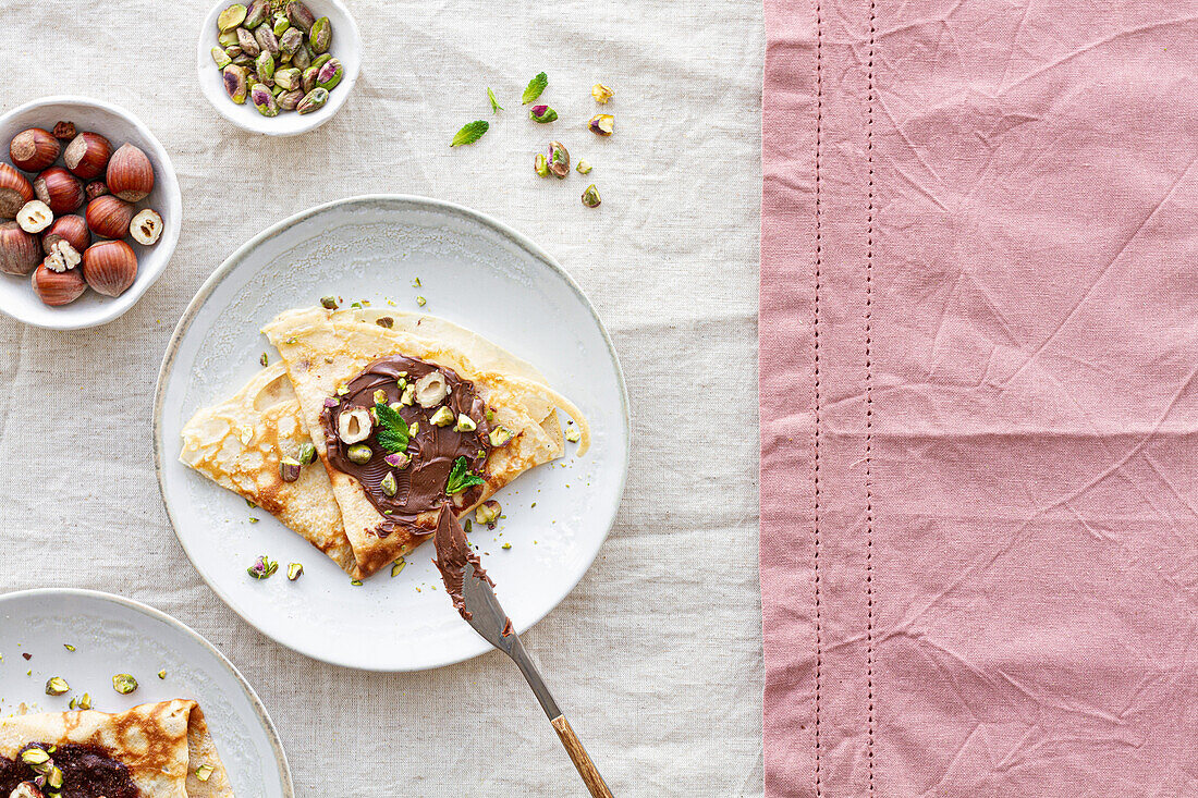 Draufsicht auf leckere Crêpes, die mit Schokolade und Nüssen garniert auf einem Teller auf dem Frühstückstisch serviert werden