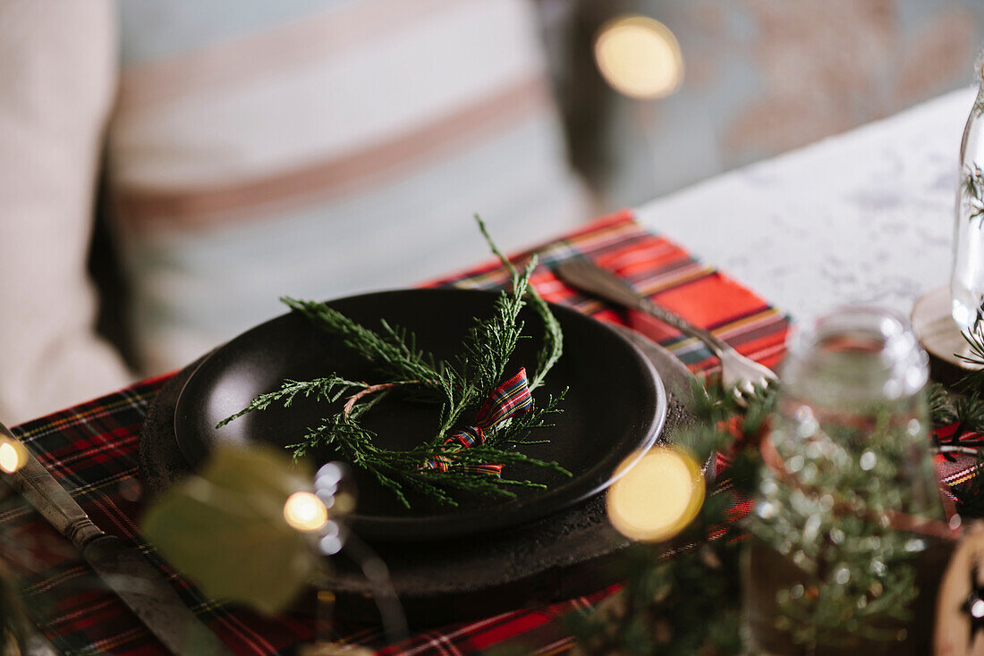 Weihnachtlich gedeckter Tisch mit Kranz auf Keramikteller auf rot kariertem Tischtuch im Hintergrund mit Lichtern