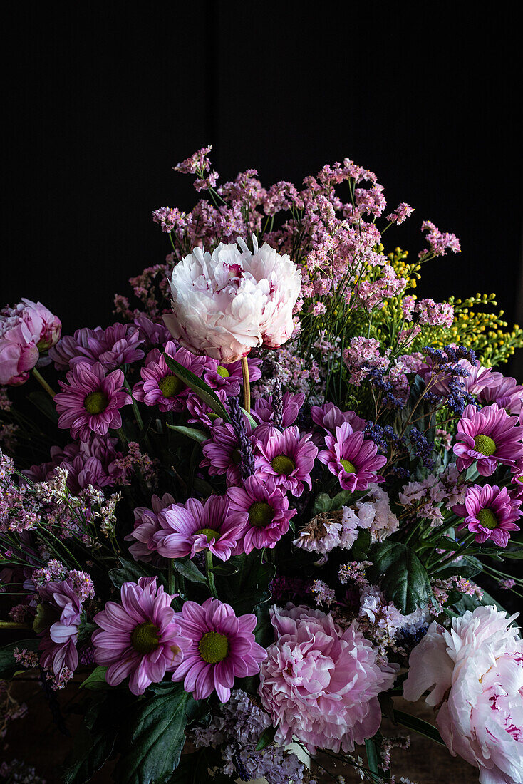 Bouquet of fresh colorful peonies and chrysanthemums in glass vase placed on wooden table in dark room