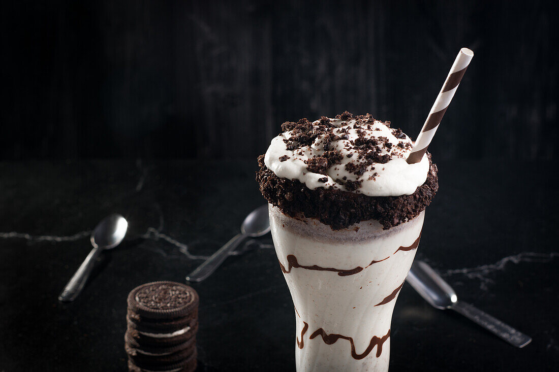 Leckerer Milchshake mit zerkleinerten Keksen und Strohhalm im Glas mit Schokoladensoße