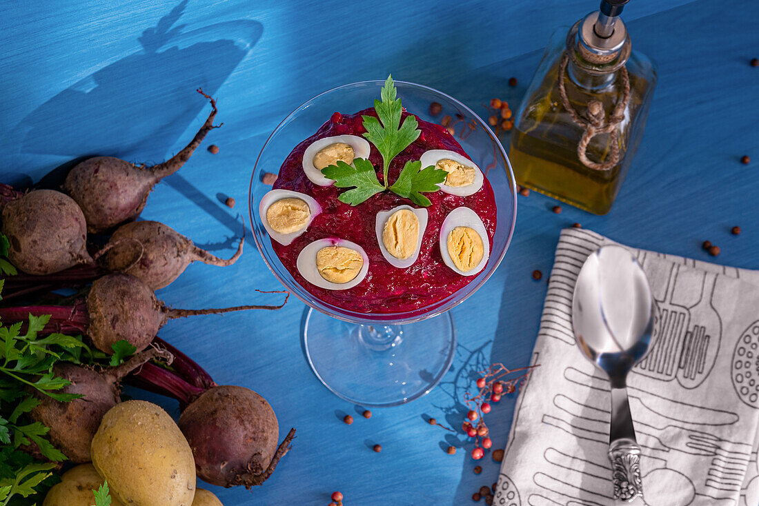 From above glass goblet of tasty beetroot cream garnished with quail eggs and parsley placed near napkin with spoon and fresh ingredients against blue background
