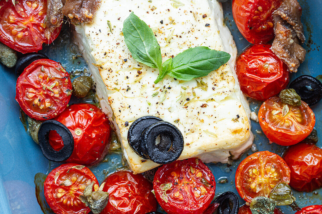 Nahaufnahme von reifen Kirschtomaten mit schwarzen Olivenscheiben und Feta-Käse in einer Auflaufform neben ungekochten Penne