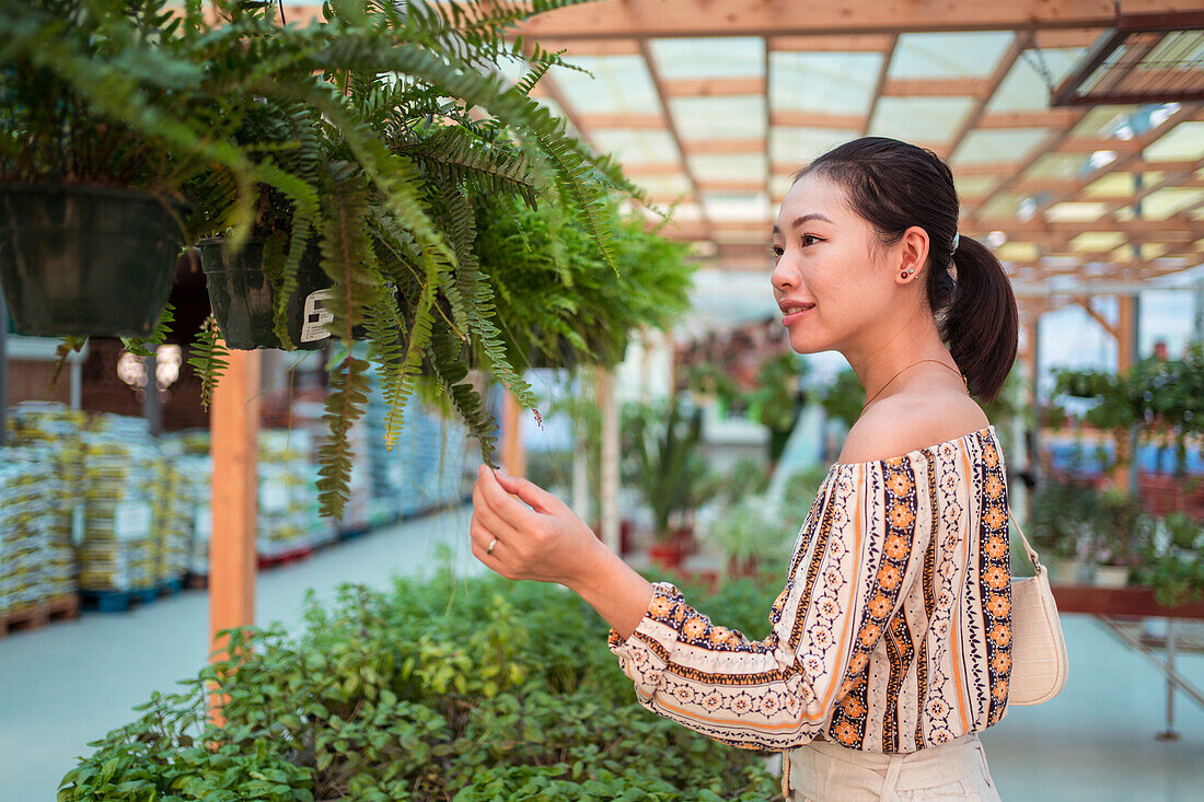 Side view of mature female shopper in textile mask picking green trees in pots in garden shop on sunny day