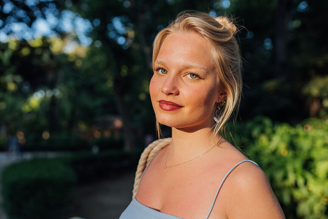 Frau mit blondem Haar steht an einem sonnigen Tag im Sommerpark und schaut in die Kamera