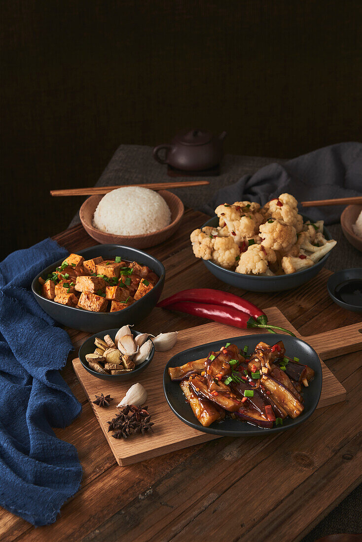 Mala-Tofu und Yuxiang, chinesische vegane Gerichte, begleitet von einer Schüssel mit Reis, Blumenkohl, Sojasauce und einer japanischen Teekanne auf einem mit Stoffen dekorierten Holztisch