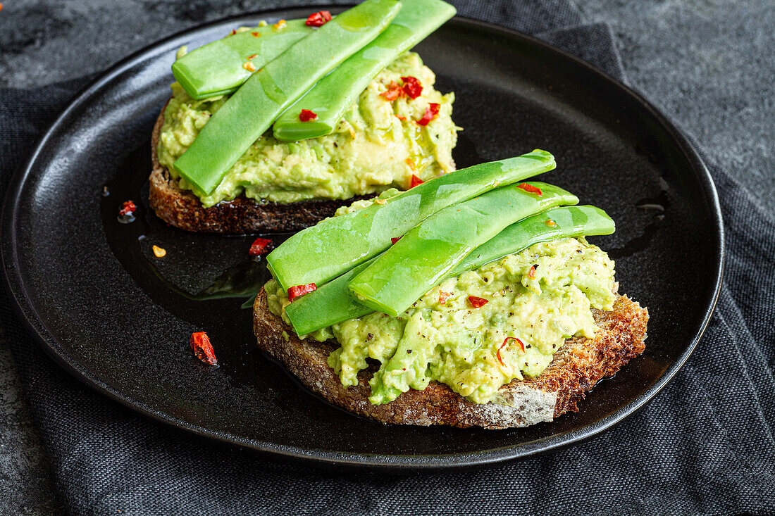 Appetitliche Toasts mit frischer Guacamole und grünen Erbsenschoten, garniert mit rotem Paprika und serviert auf einem schwarzen Teller