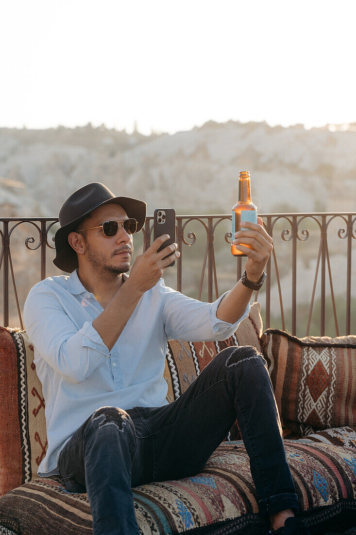 Moderner Mann in stilvollem Outfit und Sonnenbrille sitzt auf Kissen und fotografiert eine Glasflasche Bier mit dem Handy auf einer Terrassenbar in Kappadokien, Türkei