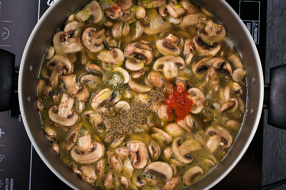 Draufsicht auf appetitlich geschnittene Pilze, die in einer Metallpfanne mit verschiedenen Gewürzen auf dem Herd während der Suppenzubereitung in der Küche kochen