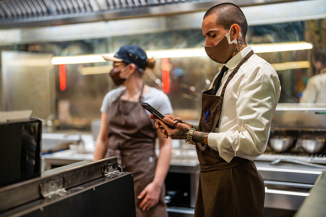 Männlicher Koch bei der Zubereitung von Speisen an der Theke in einer modernen offenen Küche neben einem Kellner, der ein Mobiltelefon im Restaurant während der Arbeit bei COVID Pandemie benutzt