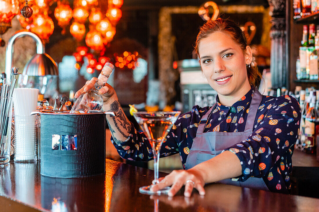 Fröhliche Barkeeperin mit einem Glas erfrischendem Wermut, die am Tresen einer Bar steht und in die Kamera schaut