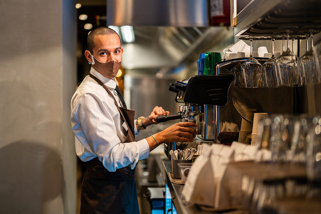 Side view of waiter in uniform and protective mask pressing buttons on coffee machine while brewing beverage in restaurant during coronavirus pandemic