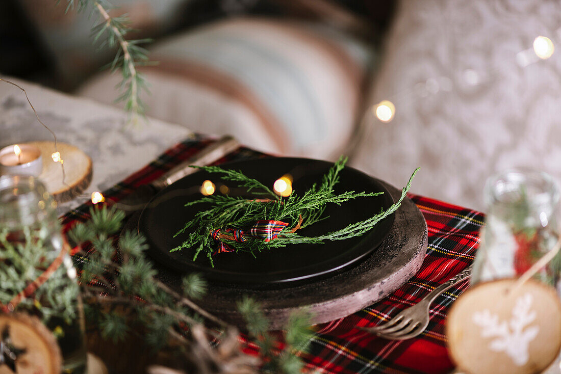 Christmas table setting with wreath on ceramic plate on red checkered tablecloth on the background with lights