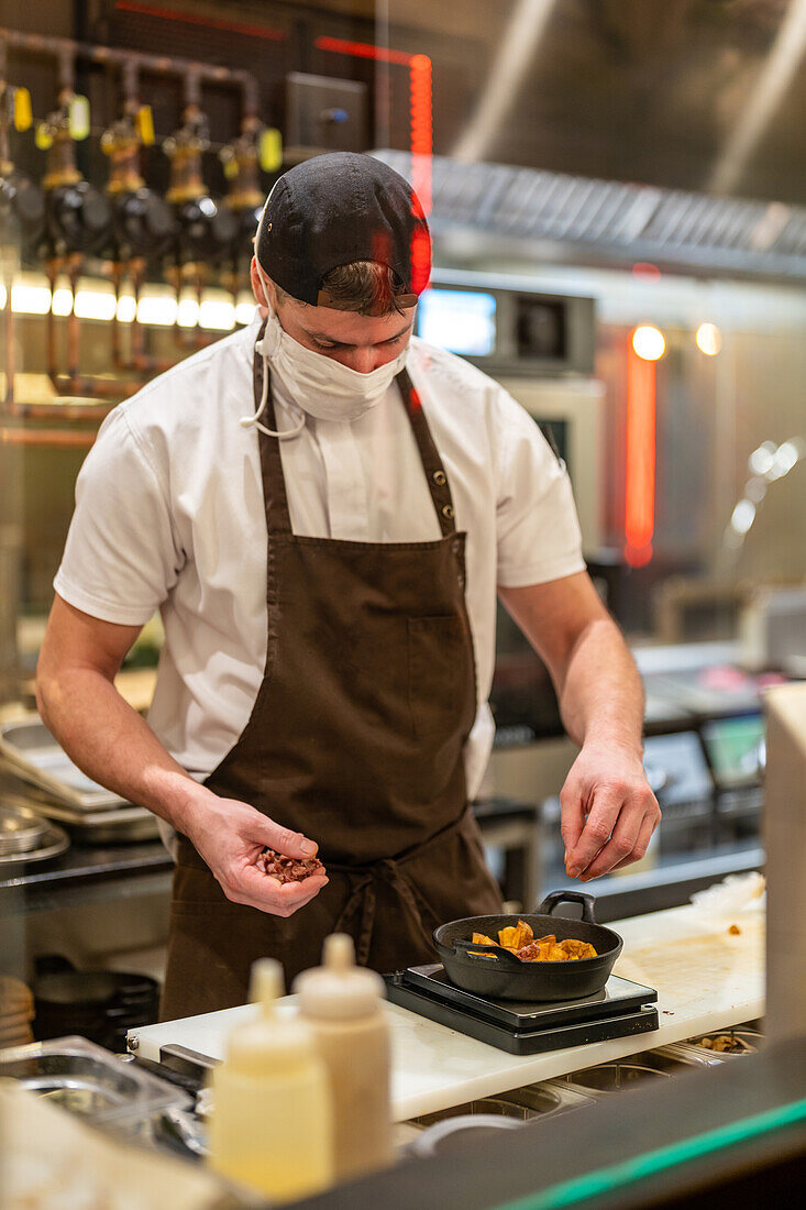 Männlicher Koch in Schürze und Mütze mit Schutzmaske, der während einer Coronavirus-Pandemie in einem Restaurant Gewürze über das Essen in der Pfanne streut