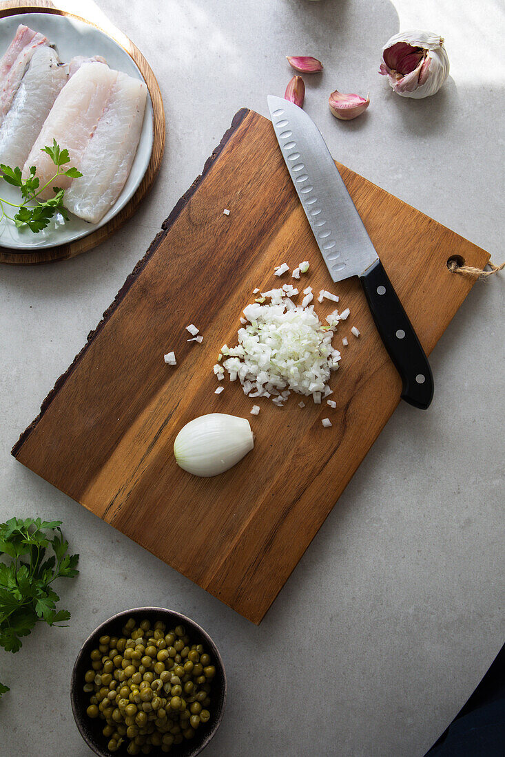 Von oben Holzbrett mit geschnittener Zwiebel und Messer neben Erbsen und Seehechtfilet mit Kräutern bei der Zubereitung von Speisen in der Küche