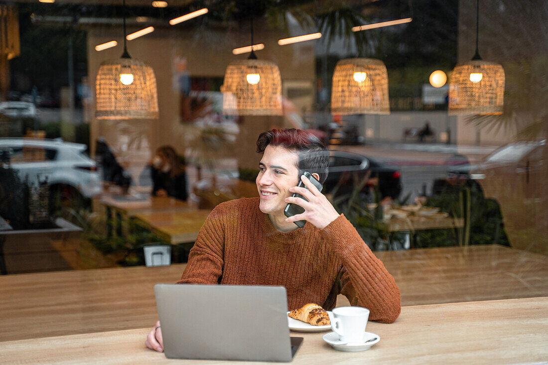 Blick durch das Fenster auf einen glücklichen jungen männlichen Studenten, der mit einem Laptop und einer Tasse Kaffee mit Croissant am Tisch sitzt und mit seinem Handy telefoniert, während er seine Zeit in einer modernen Cafeteria verbringt