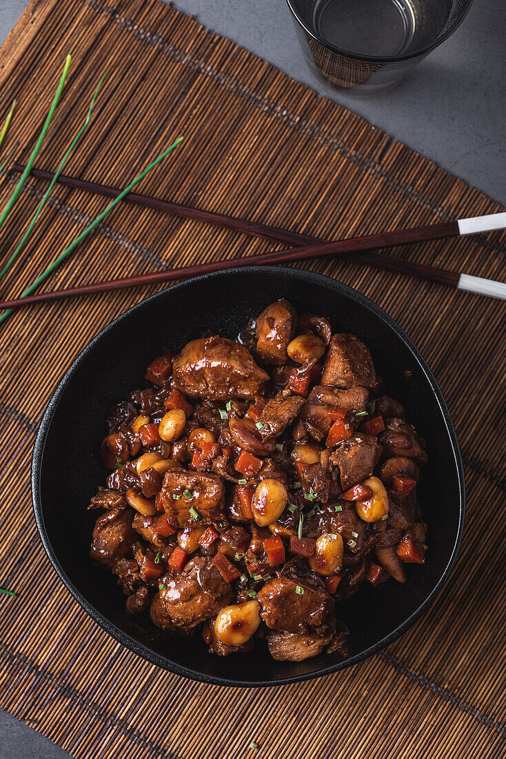 Schale mit asiatischem Hähnchenfleisch mit Mandeln und Gemüse in der Nähe von Bambus-Essstäbchen und Schnittlauch von oben