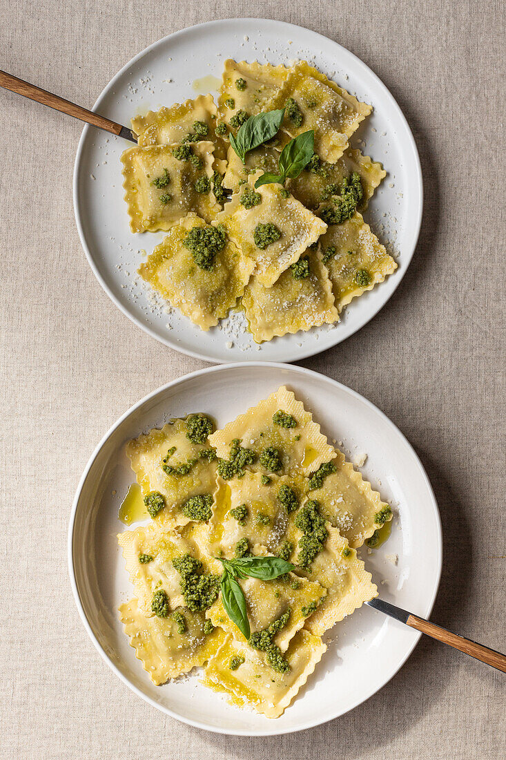 Draufsicht auf appetitlich gekochte Ravioli mit grüner Soße und Kräutern auf weißen Tellern mit Gabeln auf dem Tisch