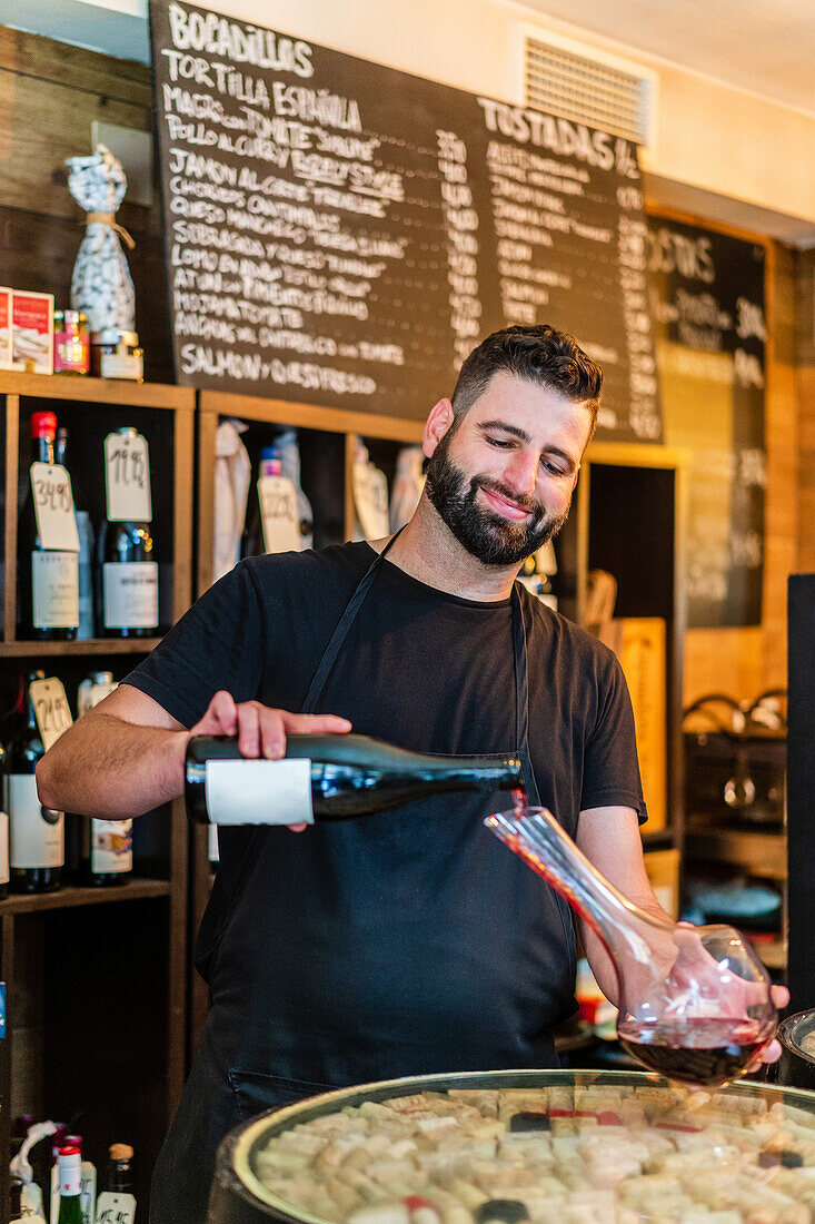 Focused man sommelier with beard and moustache in black apron standing at bar counter and pouring bottle of red wine into glass decanter in restaurant