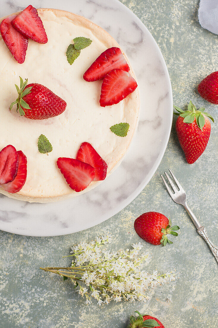 Leckerer runder Käsekuchen, dekoriert mit reifen Erdbeeren und grünen Minzblättern, serviert auf einem Teller auf einem Tisch in einer hellen Küche