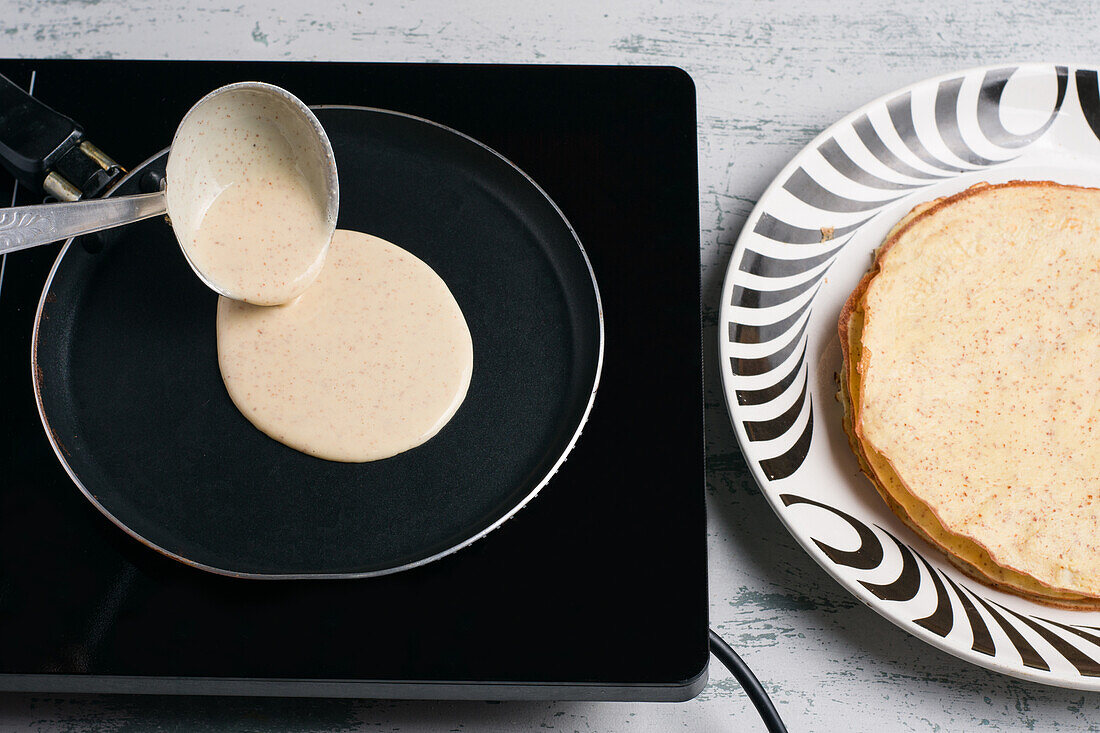 Blick von oben auf gesunden Teig für Keto-Crepe, der aus einer Kelle auf eine schwarze Bratpfanne auf dem Herd in einer hellen Küche gegossen wird