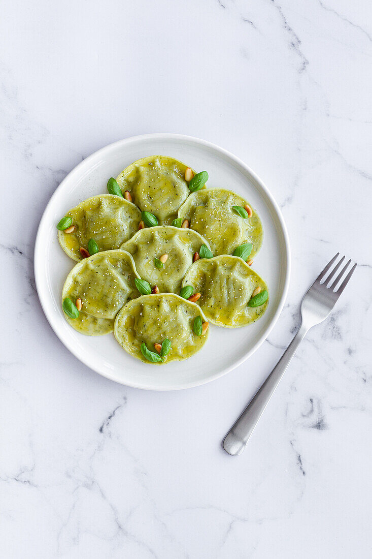 Draufsicht auf leckere Raviolis mit Basilikum und Pinienkernen auf weißem Teller in einer hellen Küche