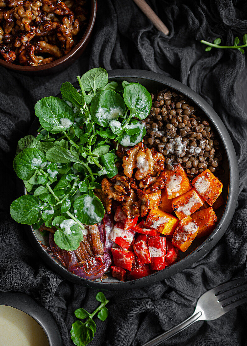 Draufsicht auf Eintopf mit Linsen neben Salat mit Kürbis und Paprika, dekoriert mit grünen Basilikumblättern auf schwarzem Hintergrund neben Salatdressing