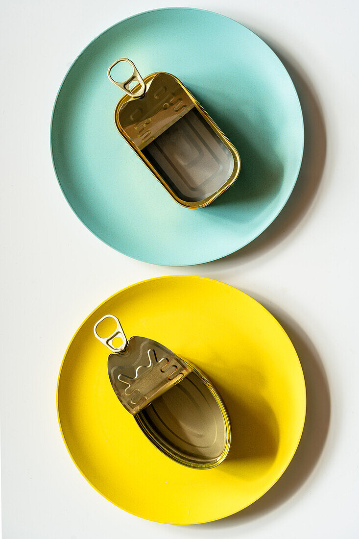 Draufsicht auf rechteckige und ovale geöffnete leere Metalldosen, die auf gelben und blauen Tellern auf einem weißen Tisch stehen