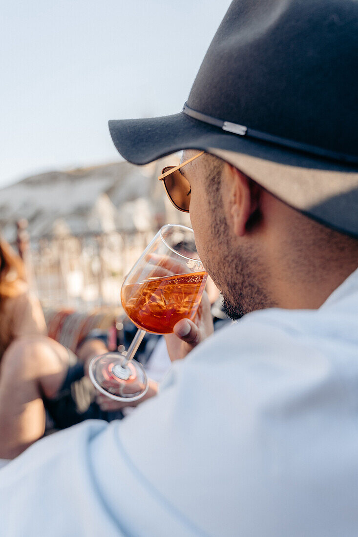 Seitenansicht eines jungen, bärtigen Mannes mit Sonnenbrille und modischem Hut, der auf der Terrasse einen kalten, erfrischenden Cocktail schlürft