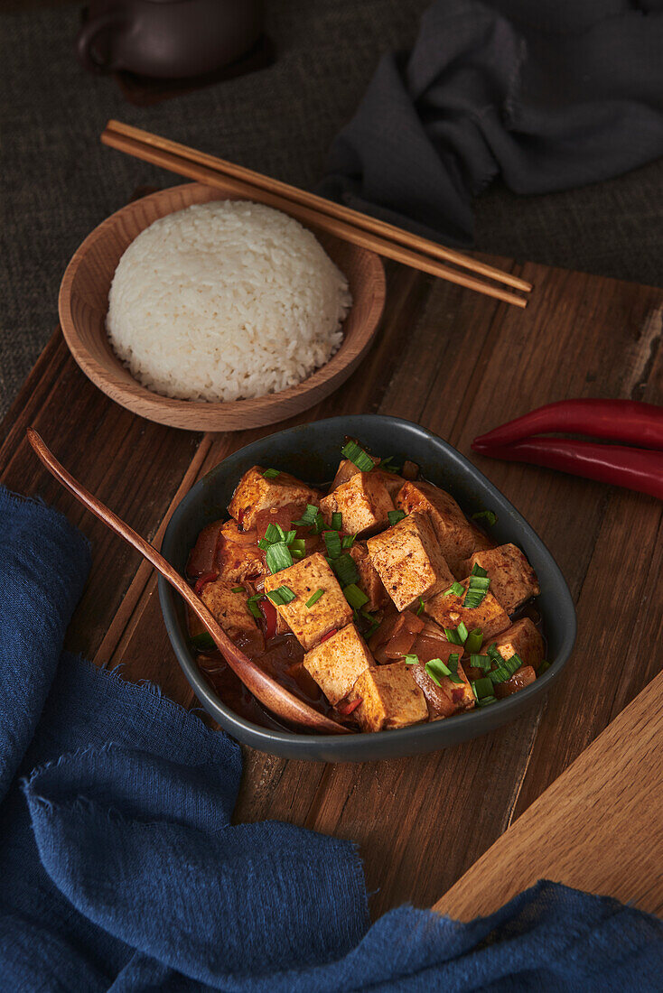 Nahaufnahme von Mala Tofu, einem chinesischen veganen Gericht, begleitet von einer Schüssel Reis und einer japanischen Teekanne auf einem mit Stoffen dekorierten Holztisch