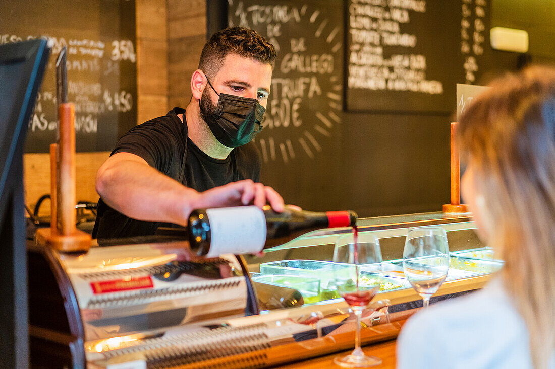 Concentrated barman in protective mask and apron standing at counter and pouring red wine into glasses for unrecognizable woman customer in cafe during coronavirus