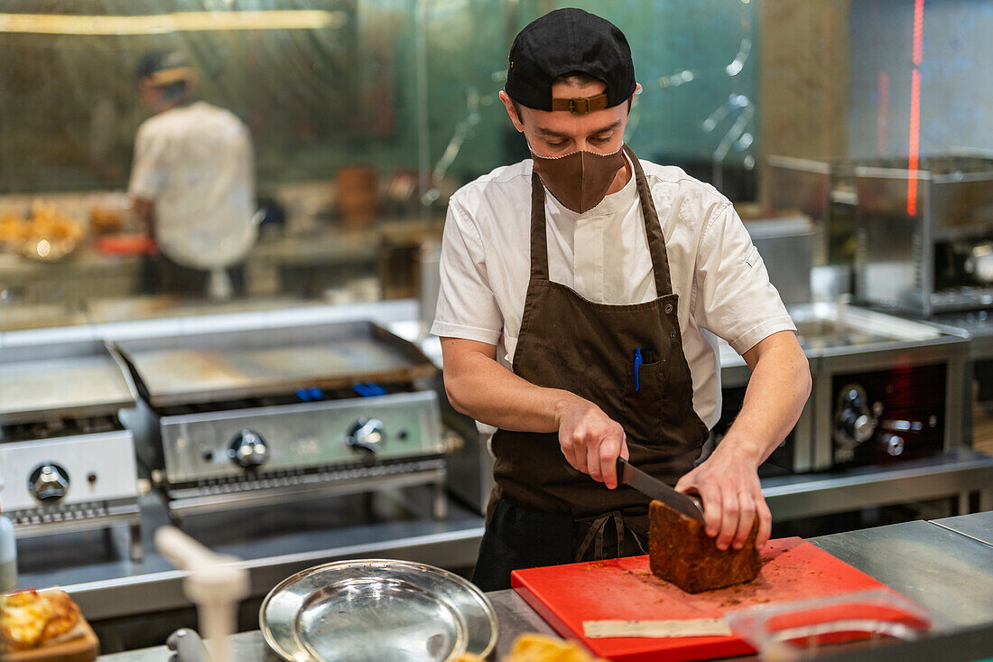 Männlicher Koch in Grau und Maske steht am Tresen und schneidet Brot auf einem Schneidebrett in einem Restaurant während einer Pandemie