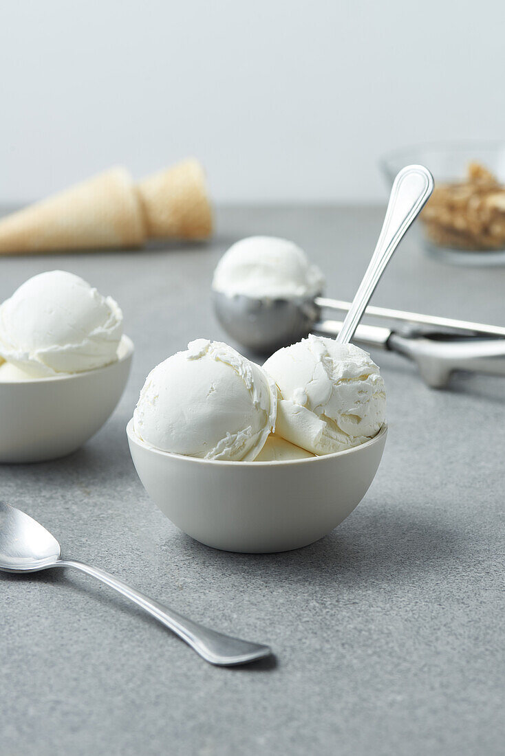 Coops of vanilla ice cream served in white ceramic bowls with spoons near waffle cones scattered on gray table